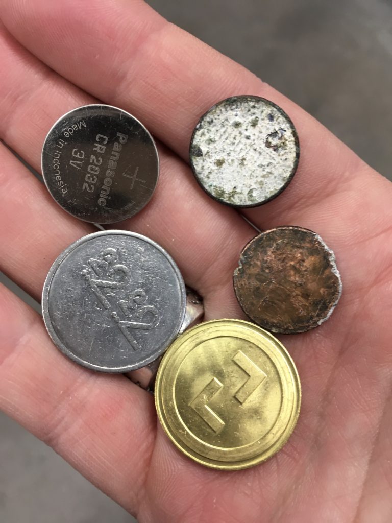 Small round objects held aloft in a hand, including two arcade tokens, a cell battery, and two mangled pennies.