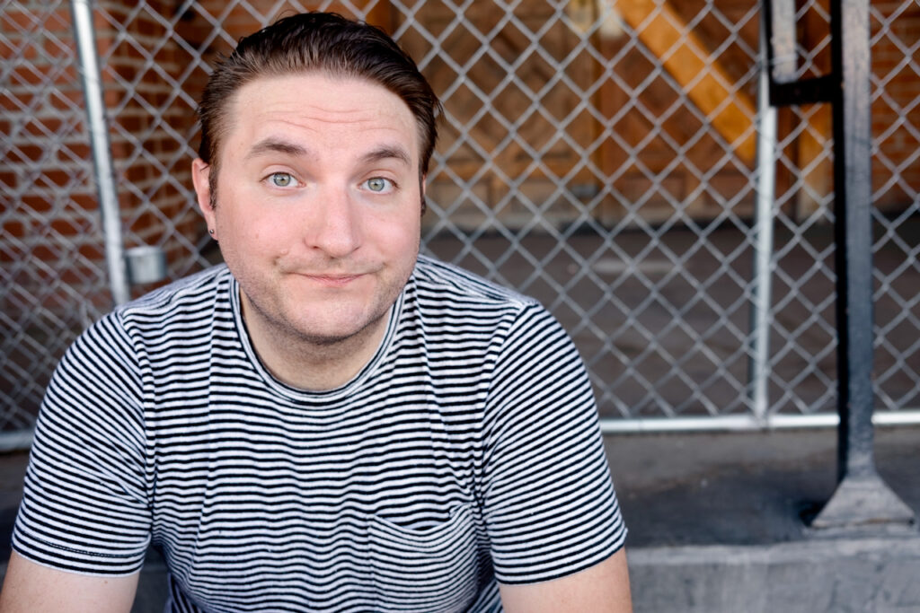 A white man with brown hair in his 30s wearing a striped shirt poses in a seated position in an urban setting in front of bricks and a chain link fence. His expression is giving "I don't know what face to make for my headshot, why does this always feel so awkward."